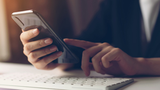 Mãos de mulher a testarem um telemóvel perto de um teclado de computador.